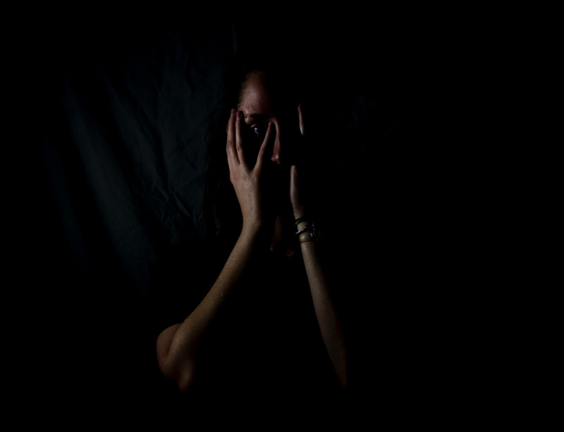 White woman with her hands in front of her face in fear, with a dark background.